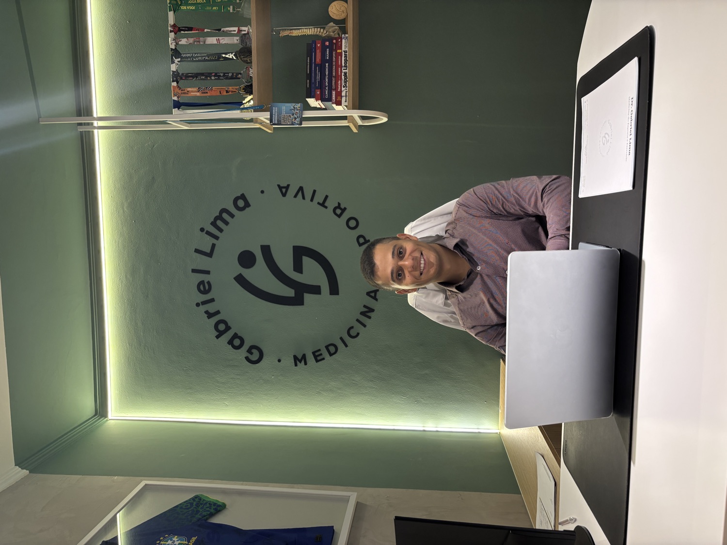 Dr. Gabriel Lima no consultório, com a marca Gabriel Lima Medicina Esportiva na parede.